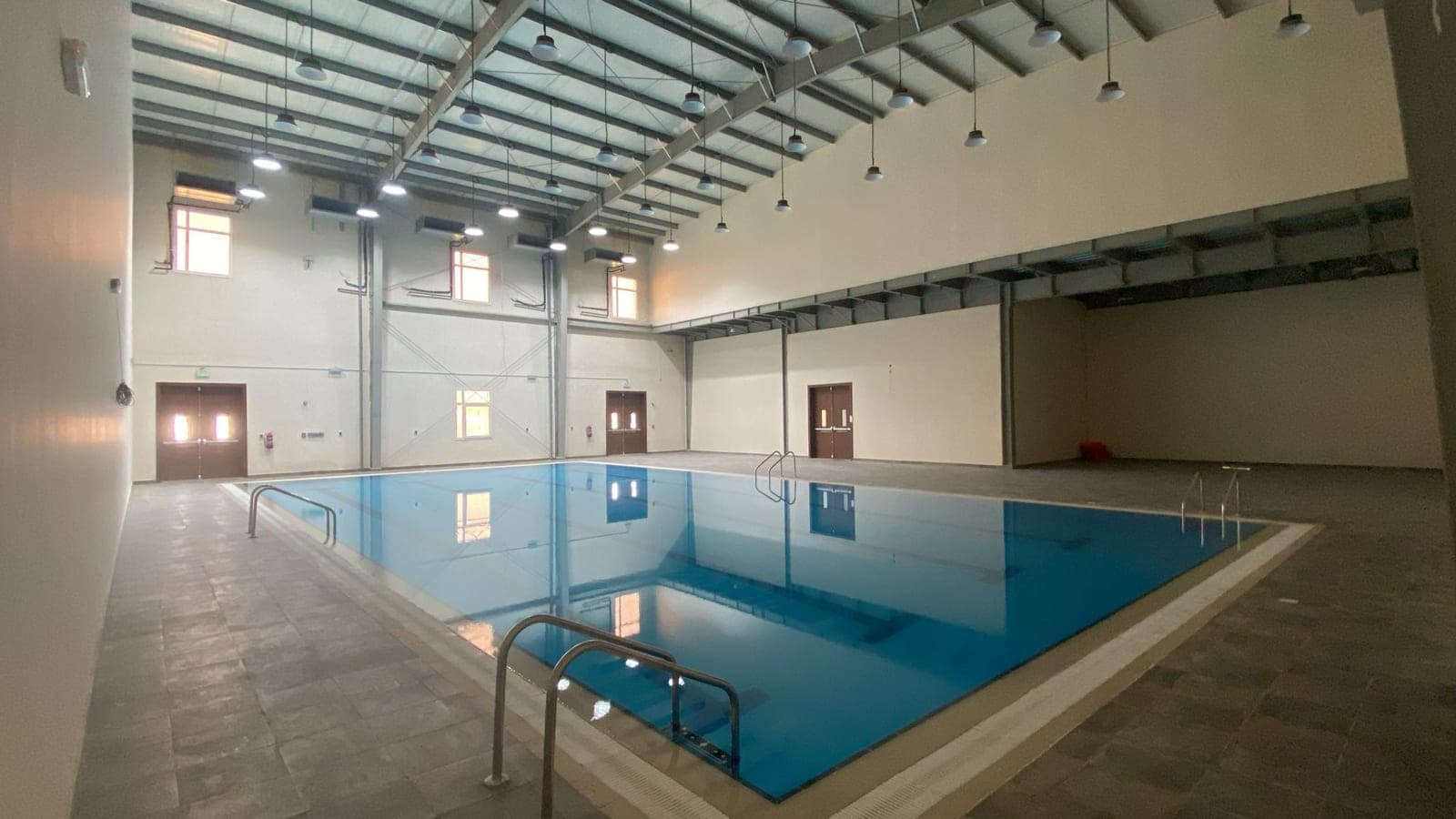 The indoor pool under its steel roof frame, clerestory windows and pendant lights above the water